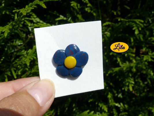 Pin’s fleur aux pétales bleu foncé avec cœur jaune réalisé en pâte polymère sur carton de présentation blanc légèrement penché vers l’arrière photographié devant une haie style floral et artisanal