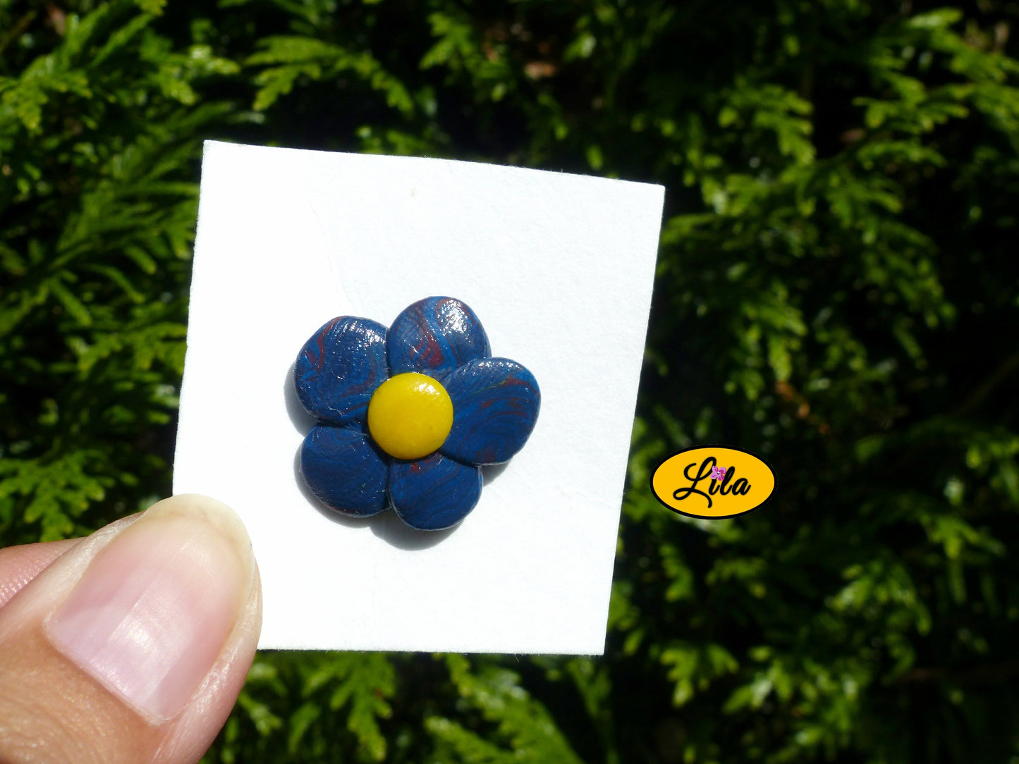 Pin’s fleur aux pétales bleu foncé avec cœur jaune réalisé en pâte polymère sur carton de présentation blanc photographié devant une haie style floral et artisanal