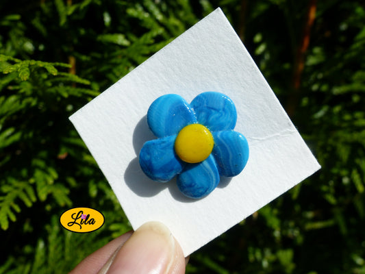 Pin’s fleur aux pétales bleus réalisé en pâte polymère sur carton de présentation blanc photographié devant une haie style floral et artisanal