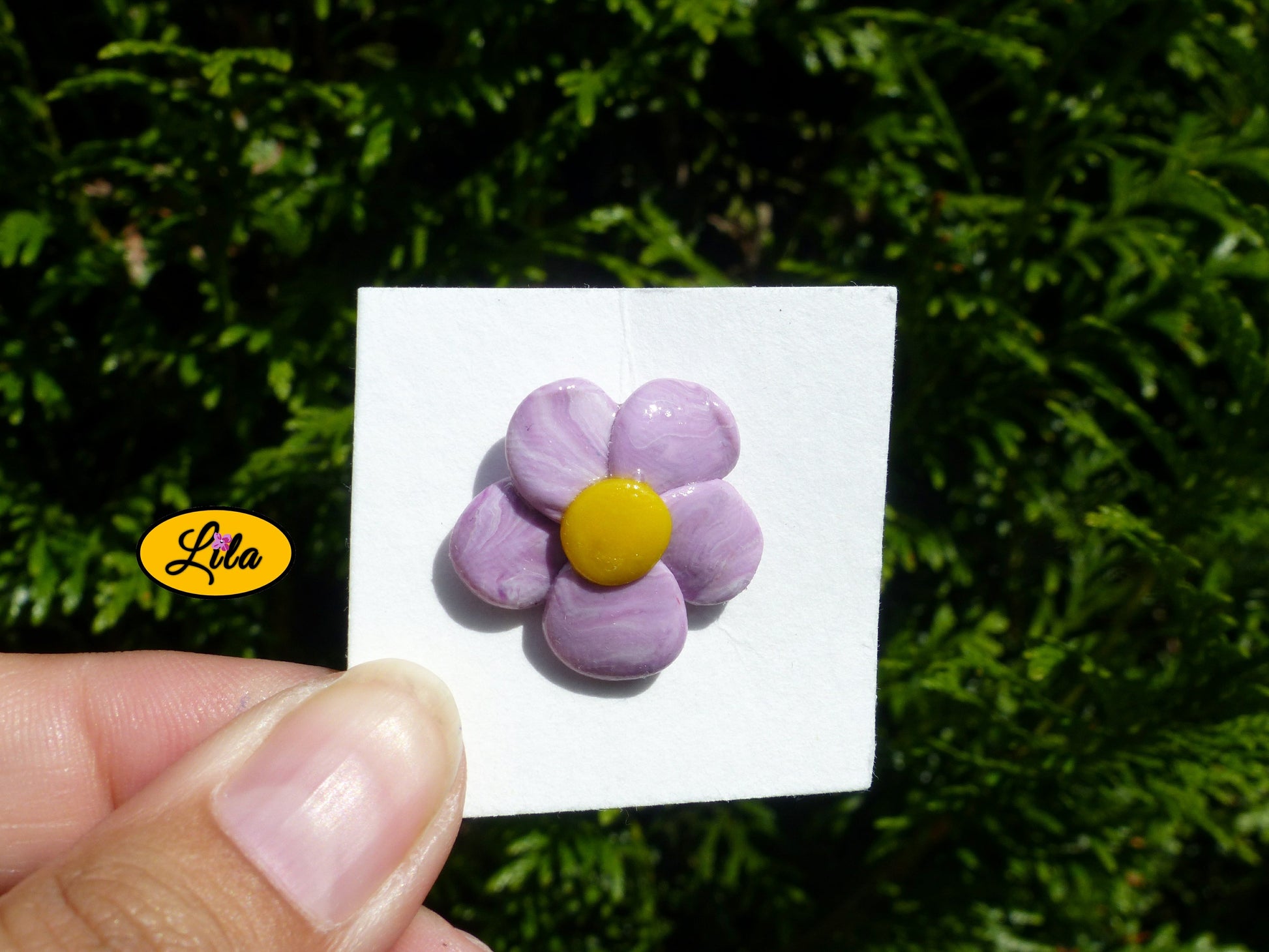 Pin’s fleur aux pétales mauves dégradés réalisé en pâte polymère sur carton de présentation blanc photographié devant une haie style floral et artisanal