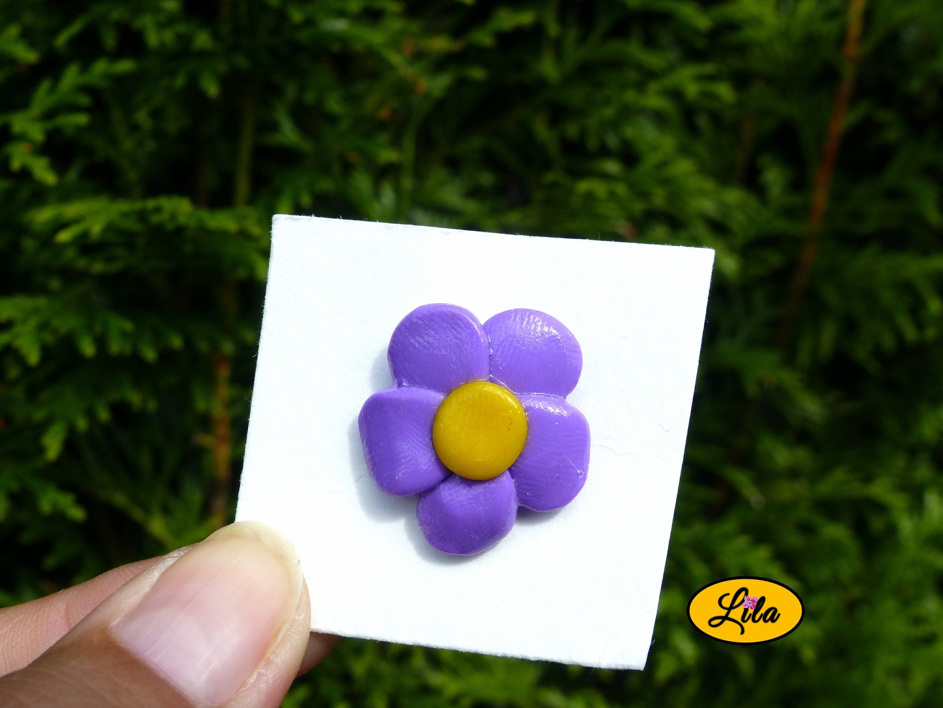 Pin’s fleur aux pétales mauves réalisé en pâte polymère sur carton de présentation blanc photographié devant une haie style floral et artisanal