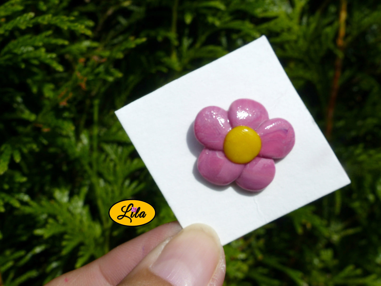 Pin’s fleur rose en pâte polymère sur carton de présentation blanc légèrement penché vers l’arrière tenu du bout des doigts devant une haie style kawaii et créatif