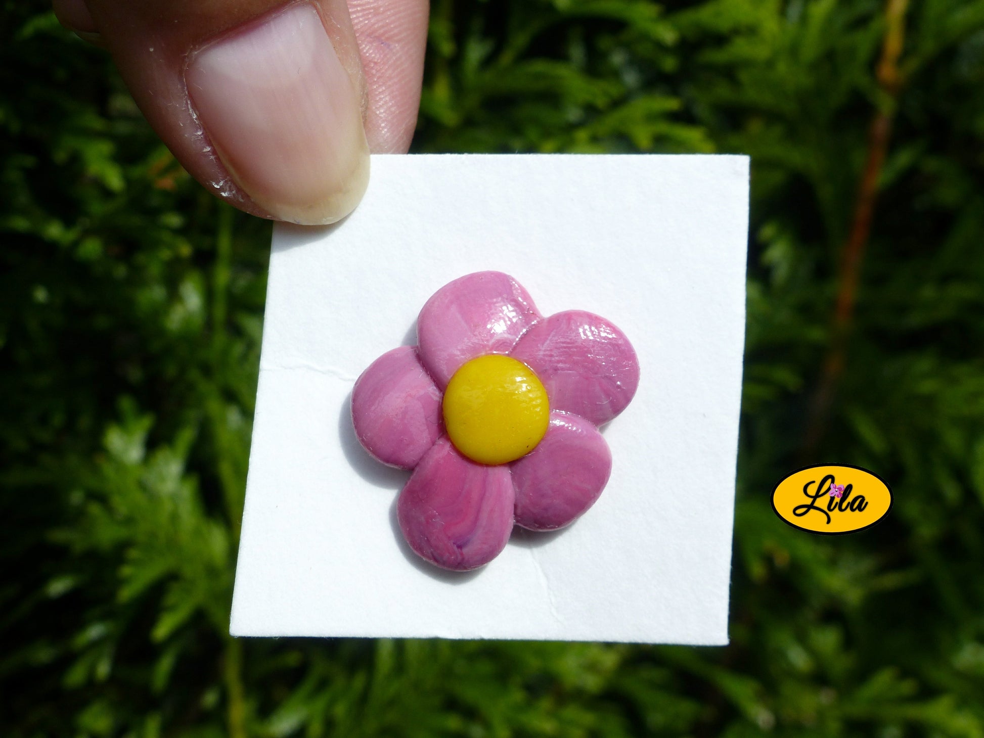 Pin’s fleur rose réalisé en pâte polymère sur carton de présentation blanc tenu du bout des doigts devant une haie style floral et artisanal