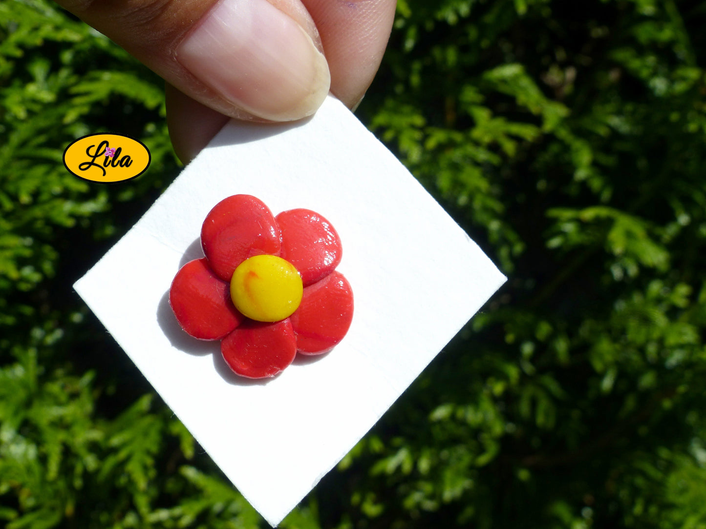 Pin’s fleur aux pétales rouges réalisé en pâte polymère sur carton de présentation blanc tourné à 180 degrés photographié devant une haie style floral et créatif