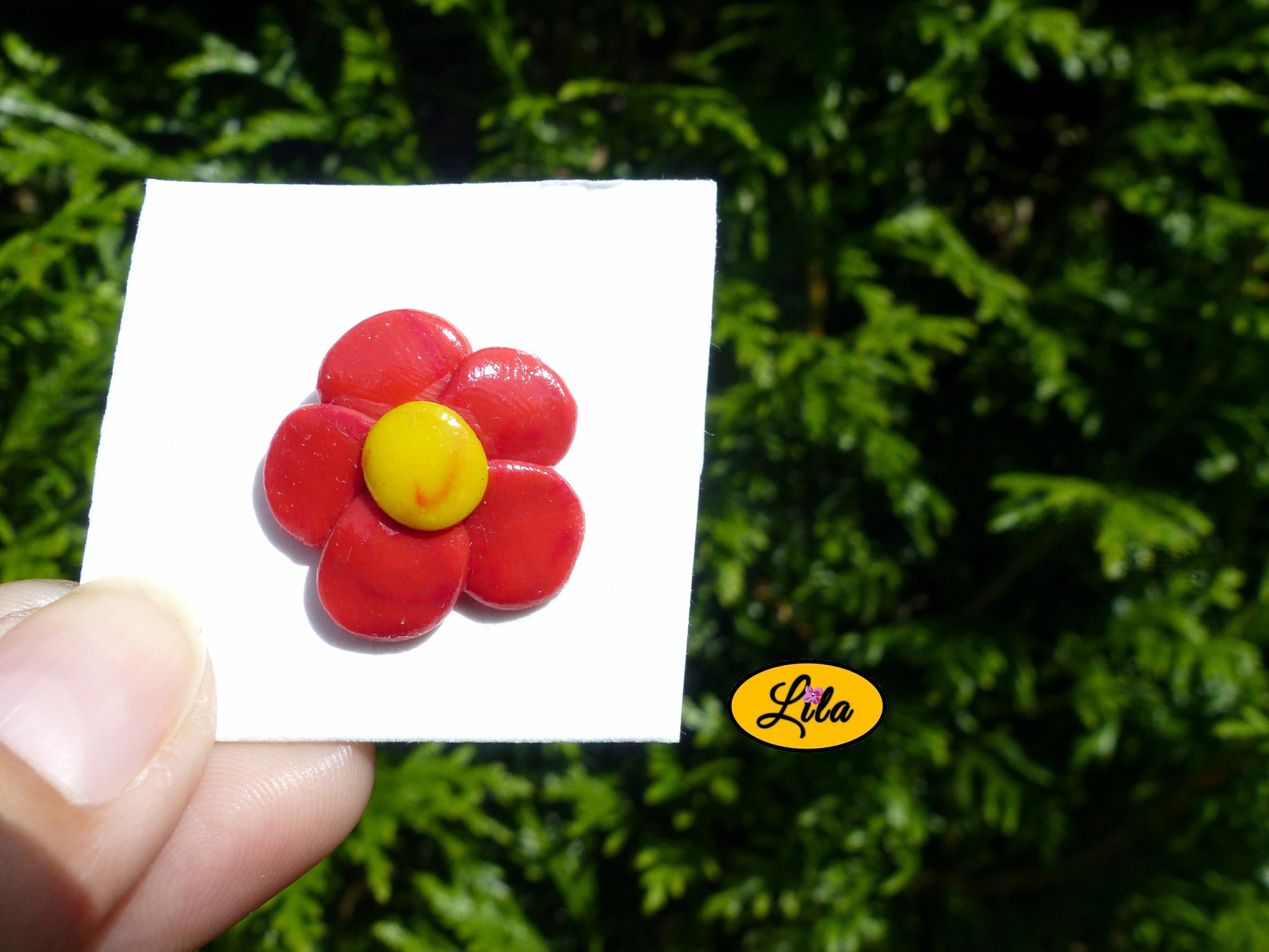 Pin’s fleur aux pétales rouges réalisé en pâte polymère sur carton de présentation blanc photographié devant une haie style floral et artisanal