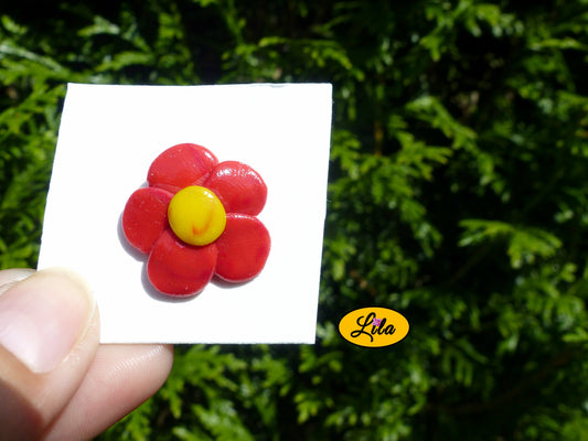 Pin’s fleur aux pétales rouges réalisé en pâte polymère sur carton de présentation blanc photographié devant une haie style floral et artisanal
