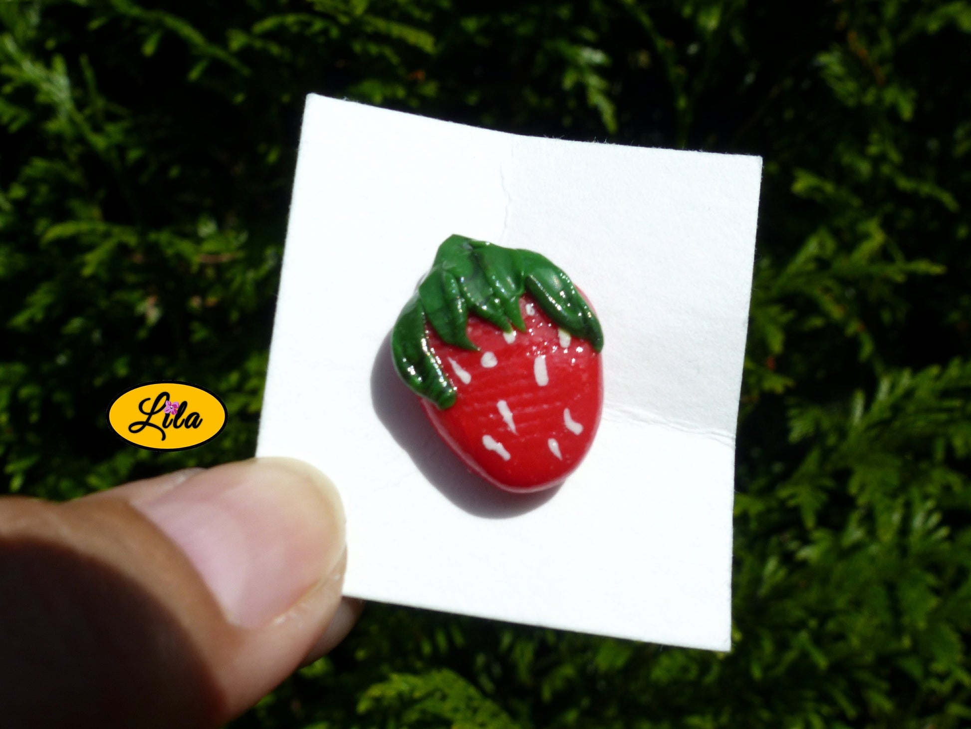 Pin’s fraise réalisé en pâte polymère sur carton de présentation blanc photographié devant une haie style gourmand et kawaii