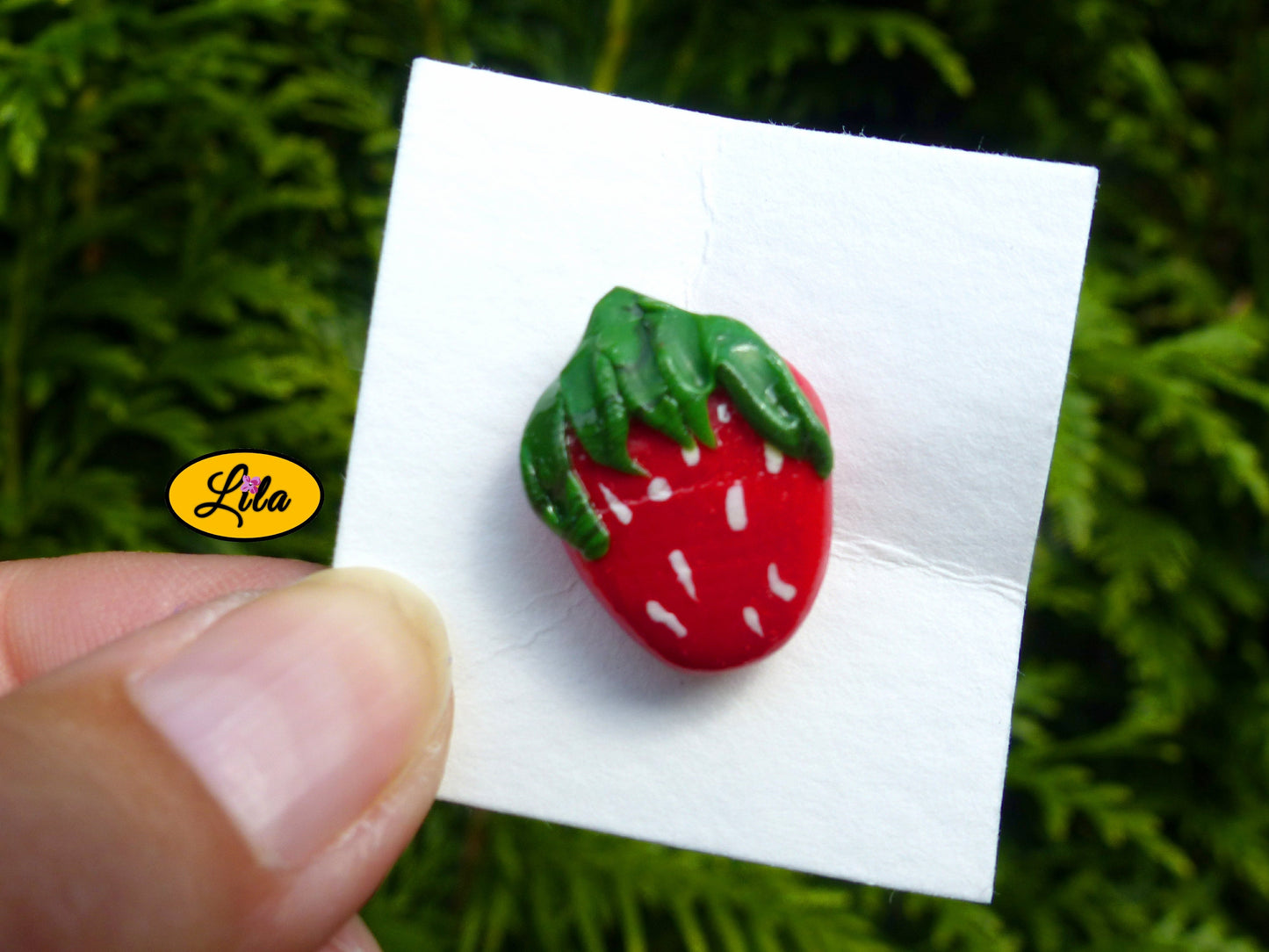 Photo rapprochée d’un pin’s fraise réalisé en pâte polymère sur carton de présentation blanc devant une haie style artisanal et gourmand