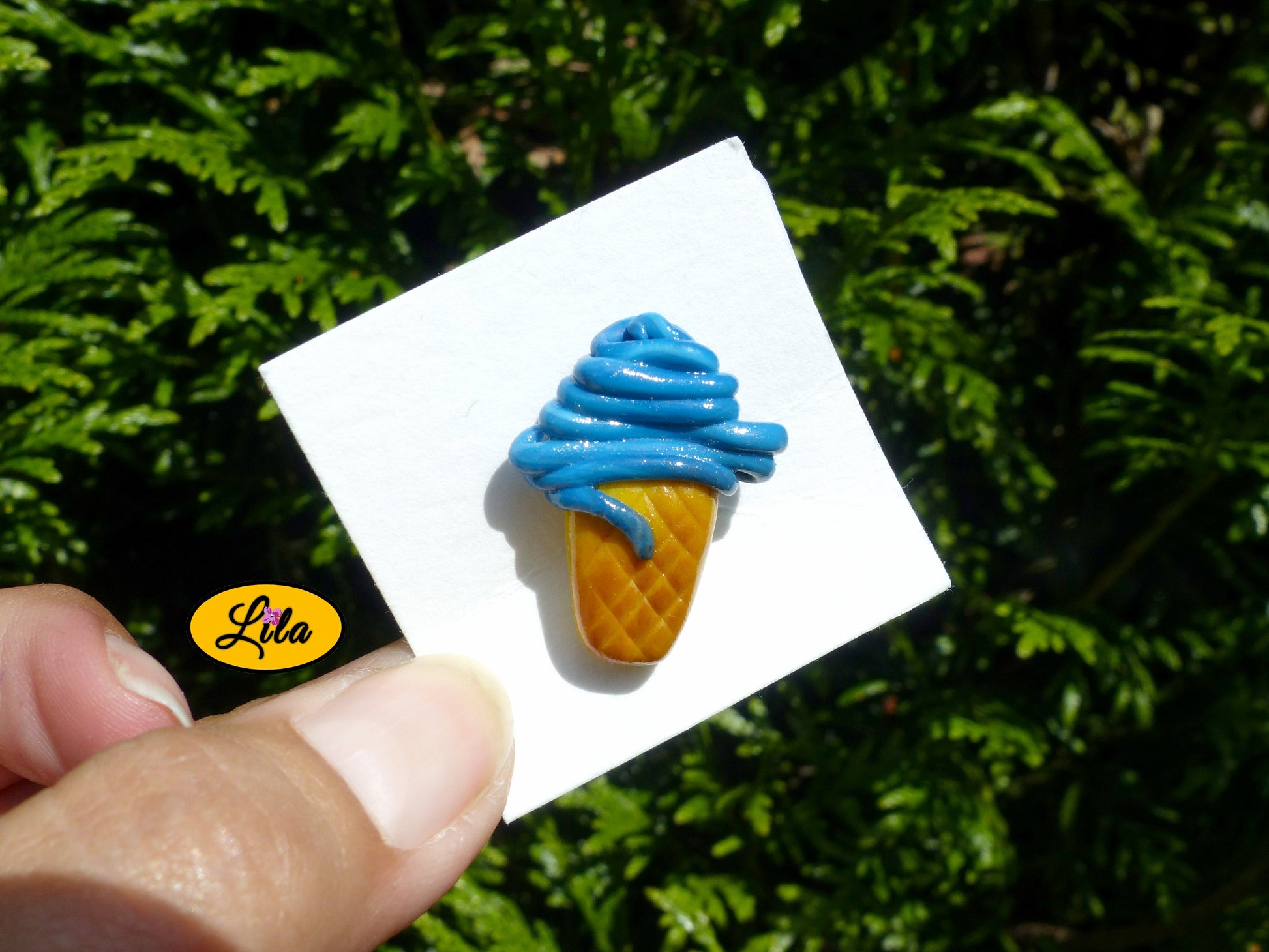 Pin’s cornet de glace italienne bleue réalisé en pâte polymère sur carton de présentation blanc photographié devant une haie style gourmand et kawaii