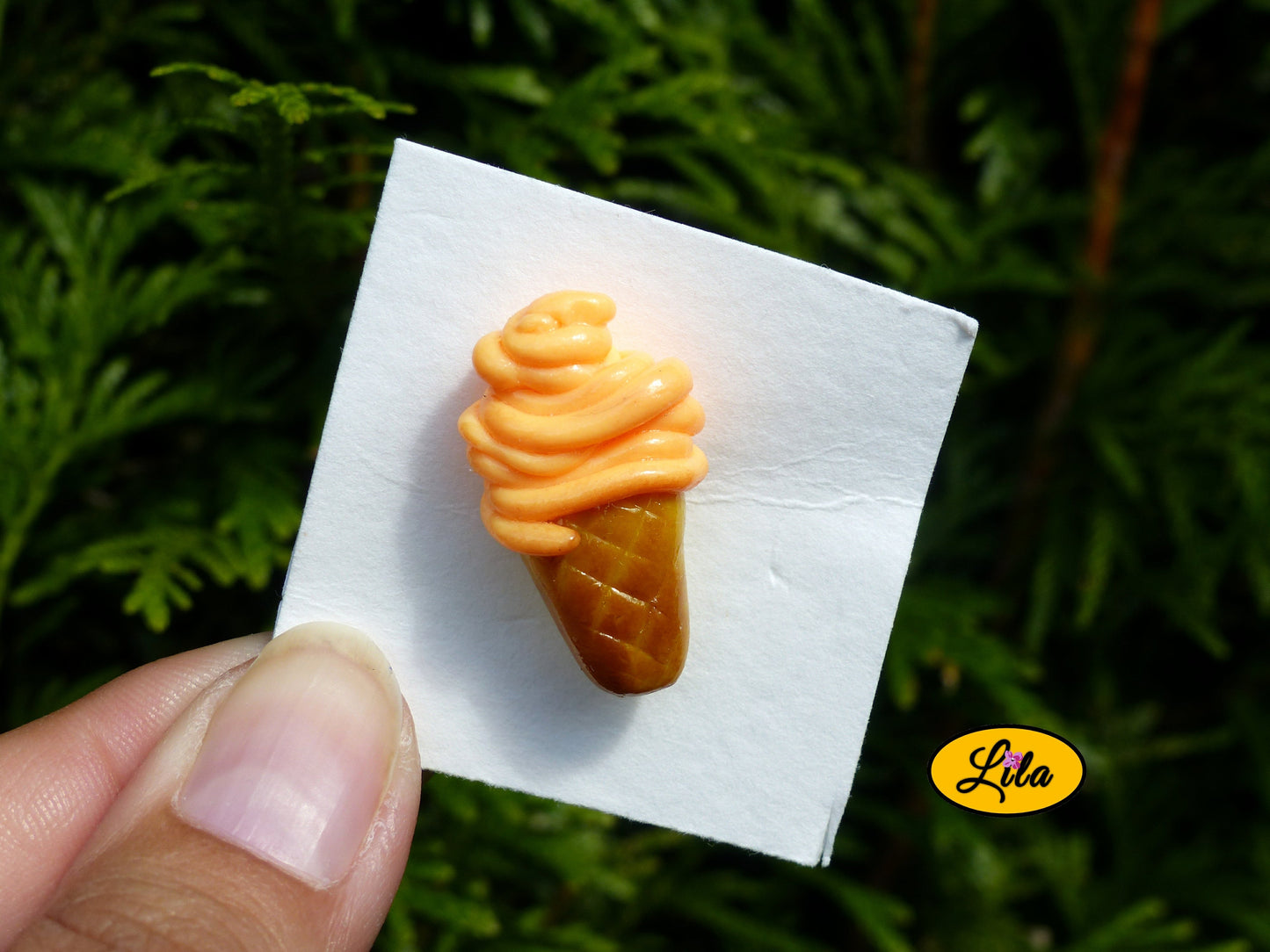 Pin’s glace orange réalisé en pâte polymère sur carton de présentation blanc photographié devant une haie style gourmand et kawaii