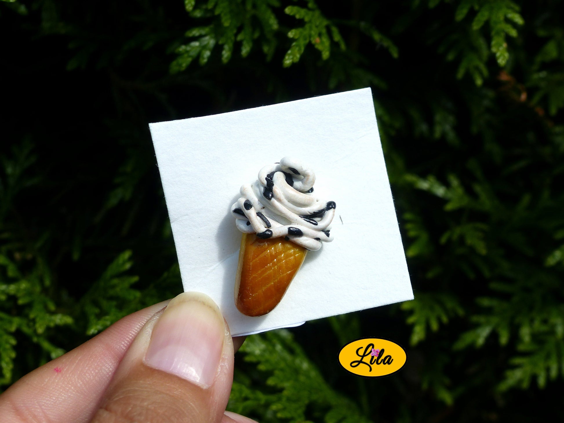 Photo rapprochée d’un pin’s glace italienne façon stracciatella réalisé en pâte polymère sur carton de présentation blanc photographié devant une haie style gourmand et kawaii