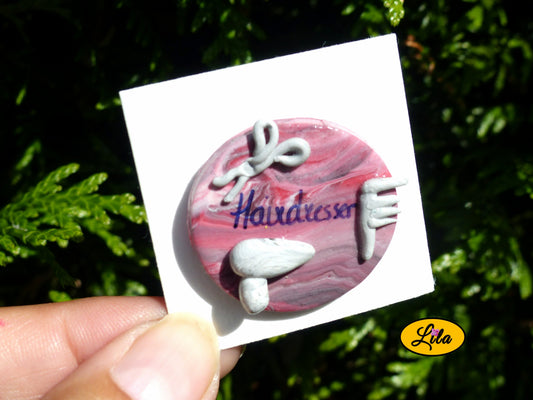Pin’s rond dégradé noir et rouge réalisé en pâte polymère avec sèche-cheveux gris, paire de ciseaux, peigne et inscription Hairdresser photographié devant une haie style cadeau pour coiffeur ou coiffeuse artisanal