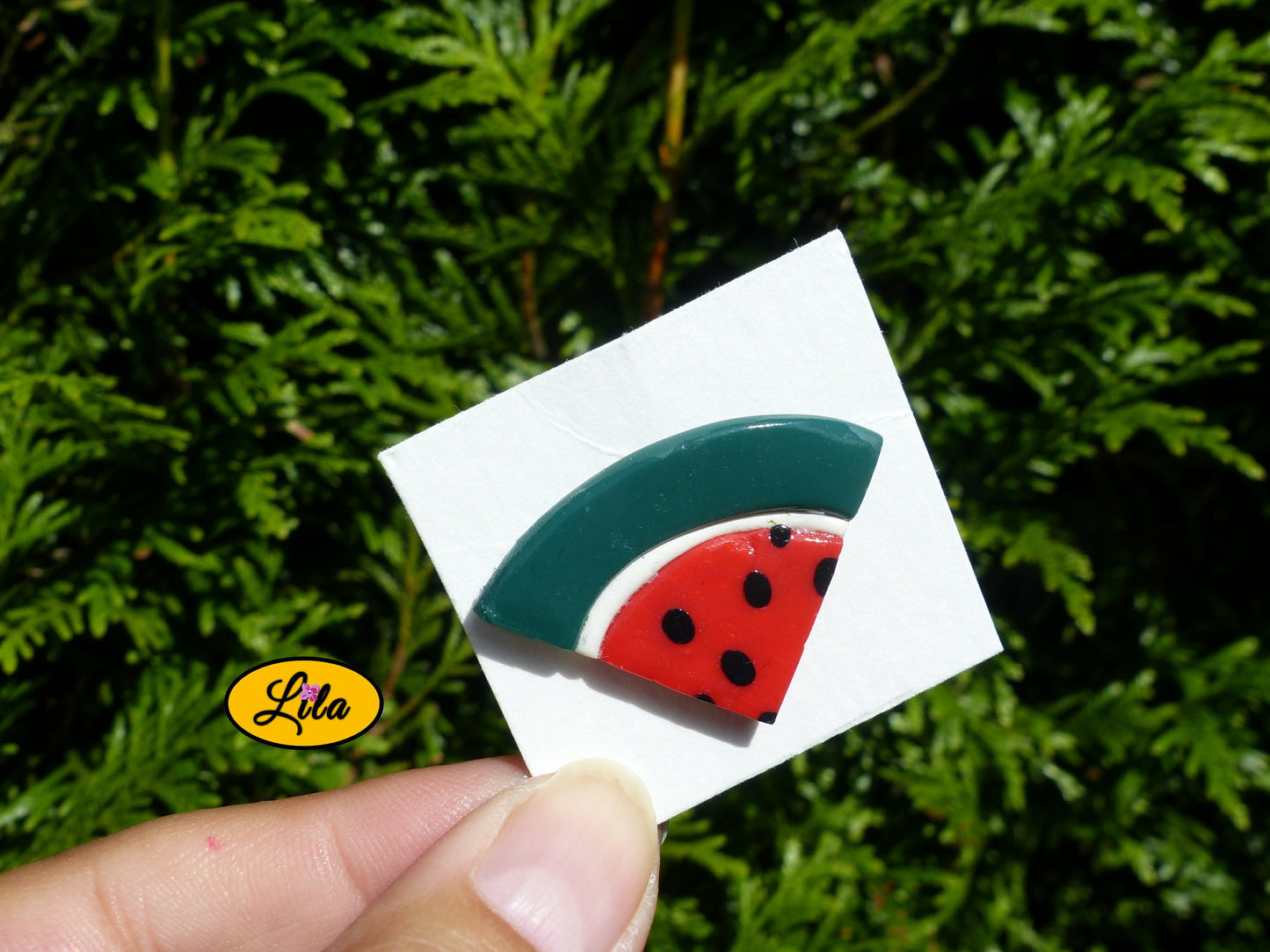 Pin’s quartier de pastèque réalisé en pâte polymère sur carton de présentation blanc photographié devant une haie style gourmand et kawaii