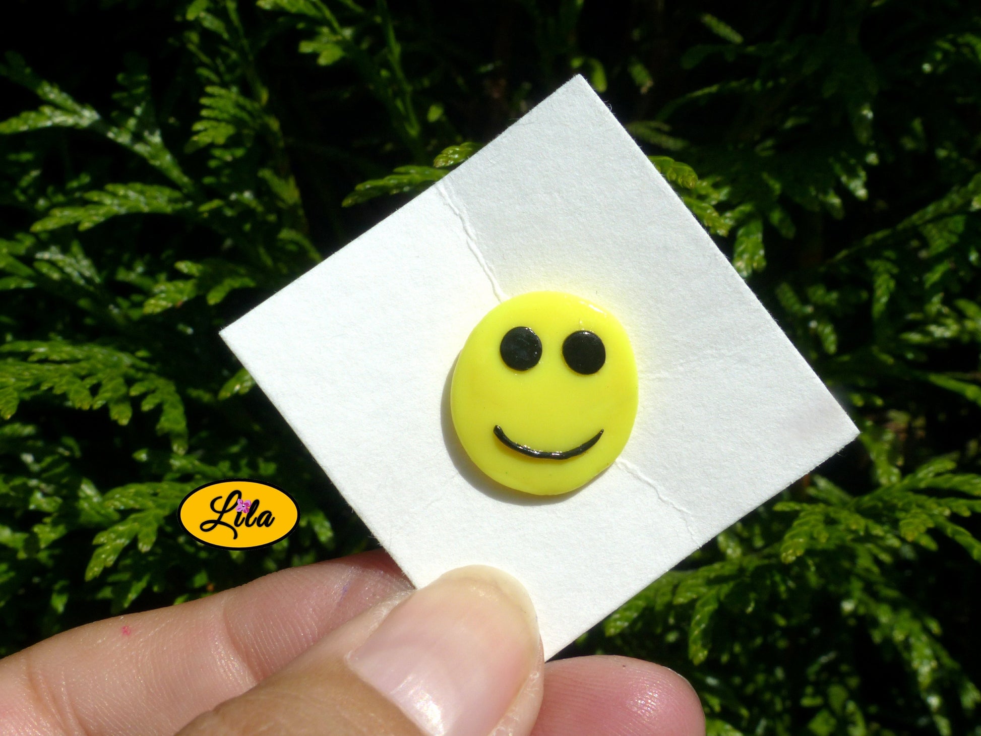 Pin’s smiley sourire réalisé en pâte polymère sur carton de présentation blanc photographié devant une haie style fun et kawaii