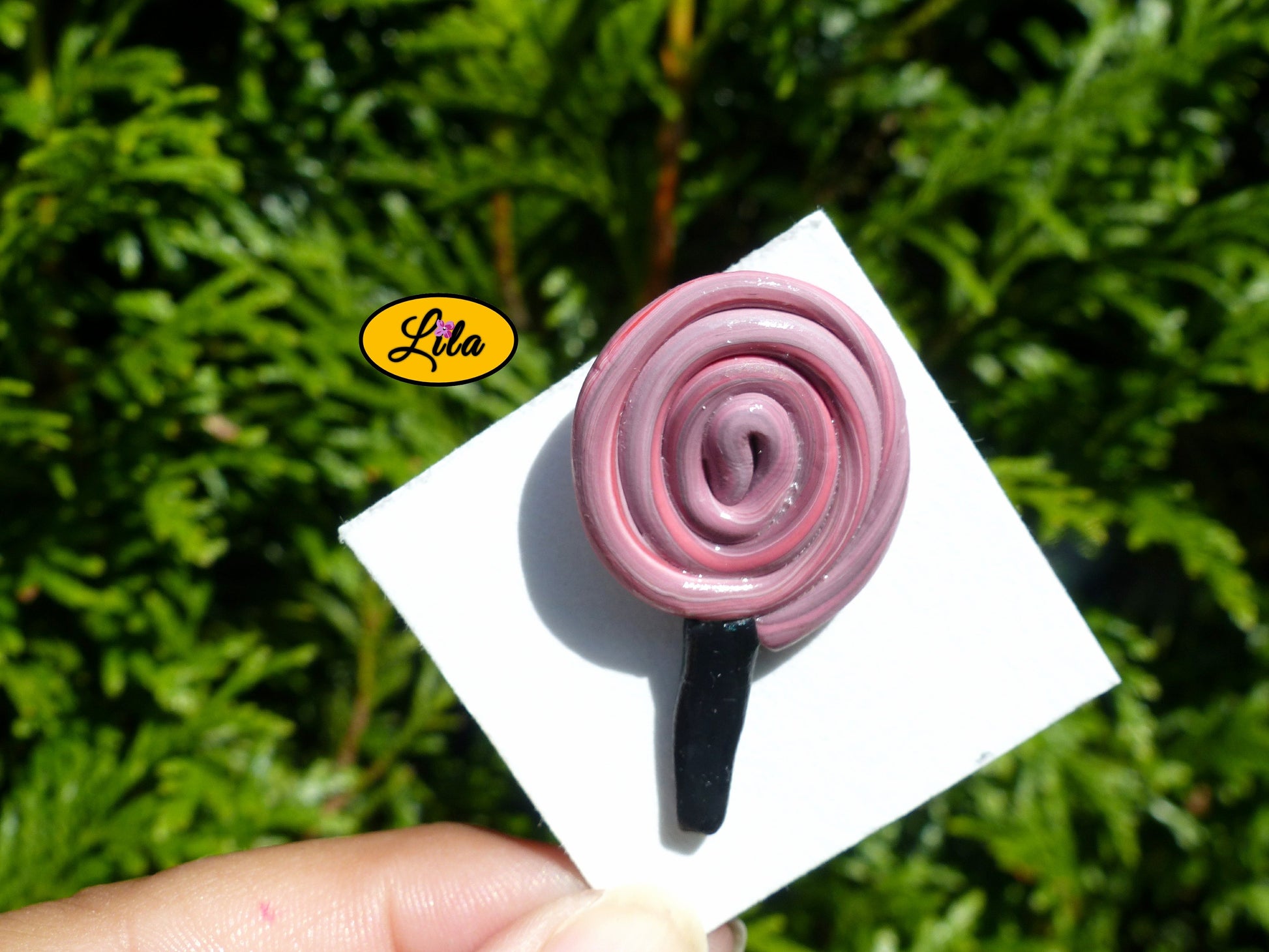 Pin’s sucette en forme de spirale rouge avec bâton noir réalisé en pâte polymère sur carton de présentation blanc photographié devant une haie style gourmand et kawaii