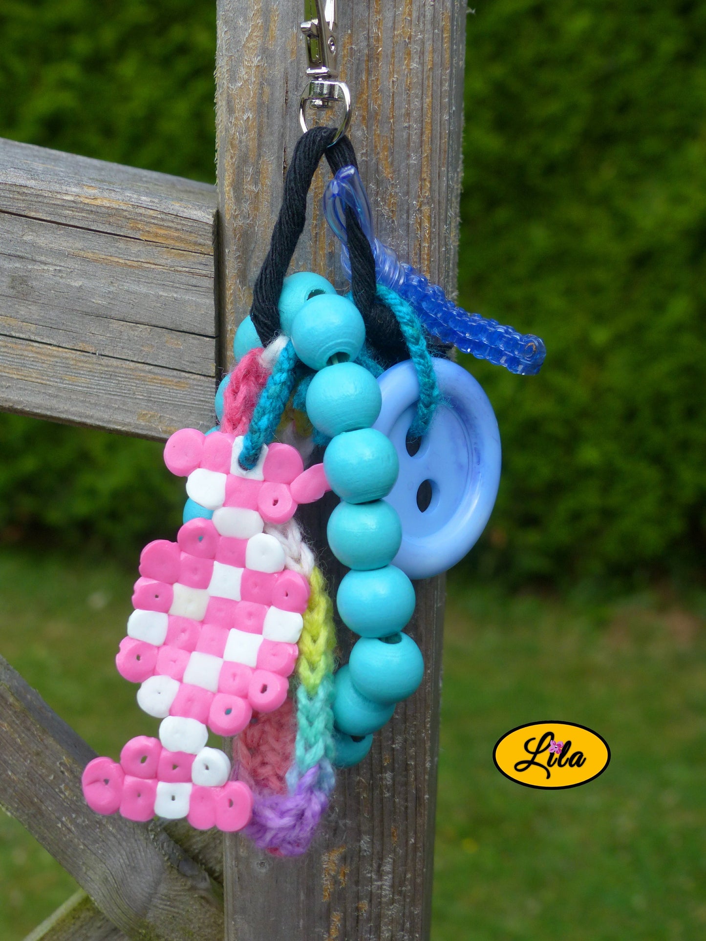 Porte-clés Bonbon Arc-en-Ciel composé d’un bonbon en perles à repasser rose et blanc, bouton bleu recyclé, languette arc-en-ciel crochetée, bracelet en grosses perles de bois bleues et mini scoubidou bleu suspendu à un mousqueton style kawaii et artisanal