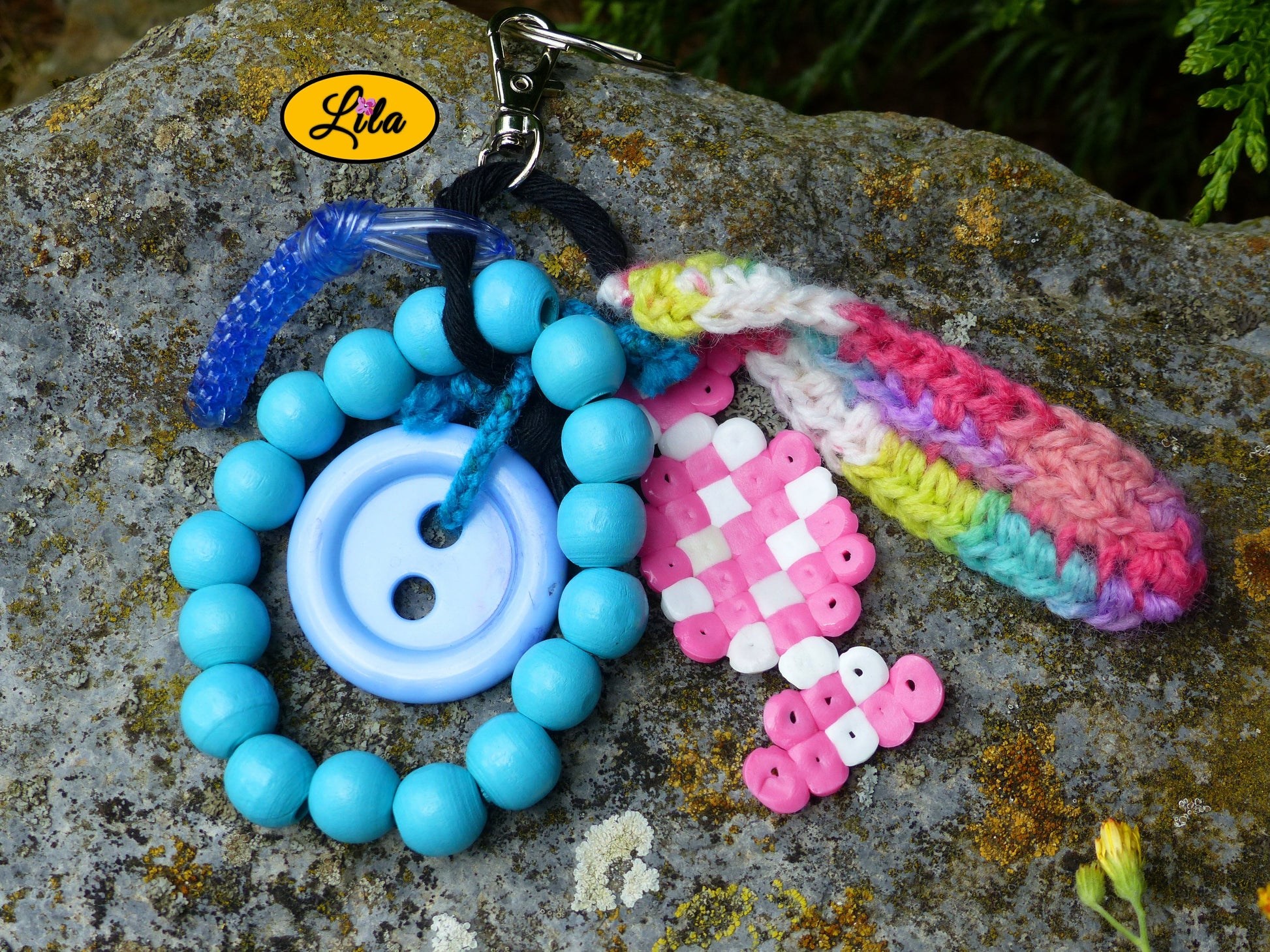 Porte-clés Bonbon Arc-en-Ciel posé sur une pierre vue de haut avec bonbon en perles à repasser rose et blanc, bouton bleu recyclé, perles en bois bleues, mini scoubidou bleu et languette crochetée arc-en-ciel visibles côte à côte style créatif et récup