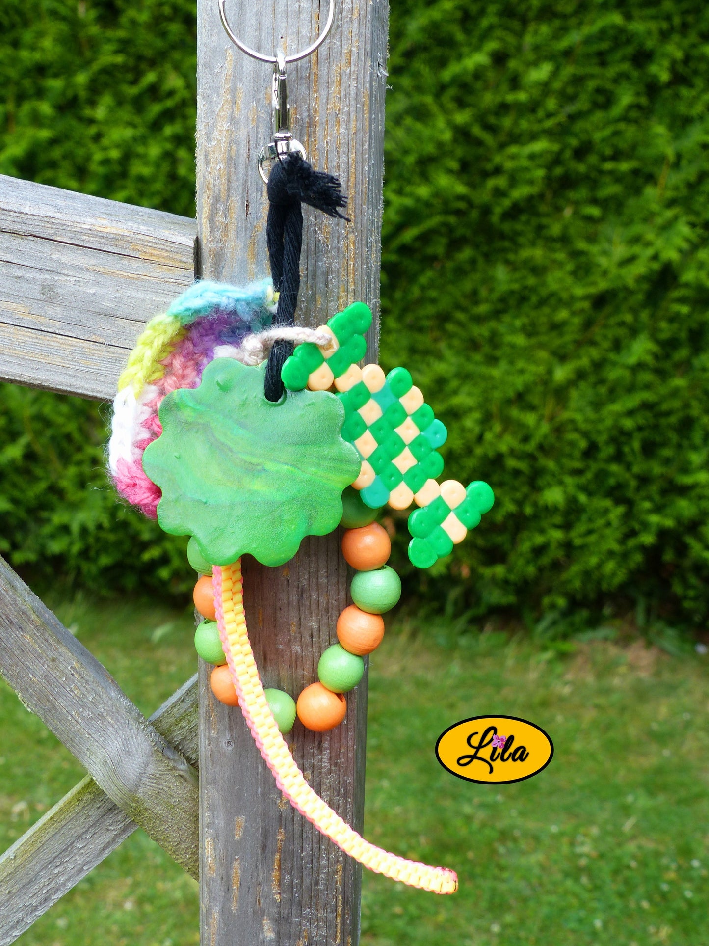 Porte-clés bonbon vert composé d’un bonbon en perles à repasser vert et orange, fleur en pâte polymère dégradé vert, perles en bois colorées, scoubidou jaune et rose et languette arc-en-ciel crochetée accroché à un morceau de bois en extérieur style kawaii et artisanal