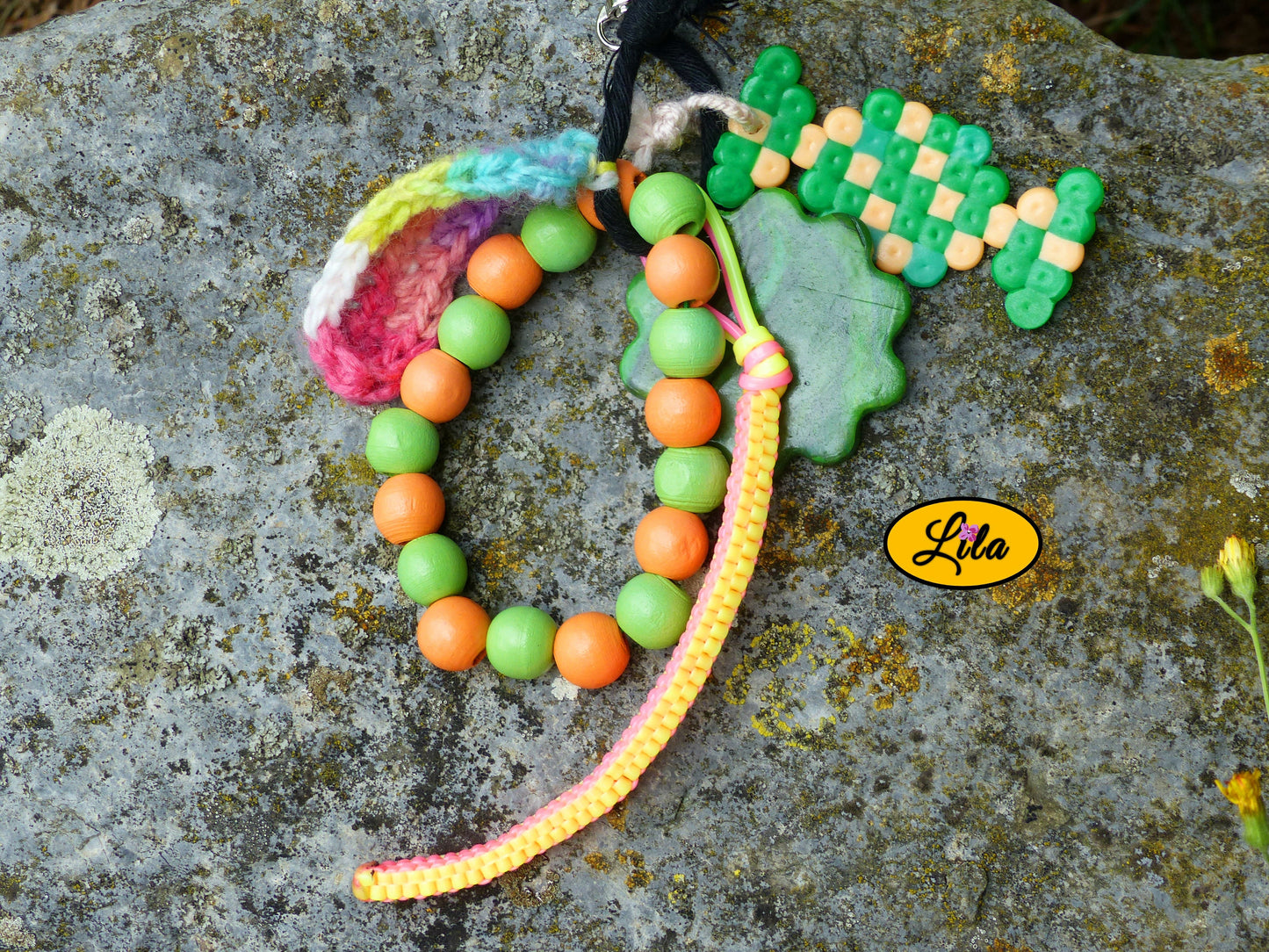 Porte-clés bonbon vert multicolore posé sur une pierre vue de haut avec bonbon en perles à repasser, fleur en pâte polymère, perles en bois, scoubidou et languette arc-en-ciel crochetée visibles côte à côte style créatif et artisanal
