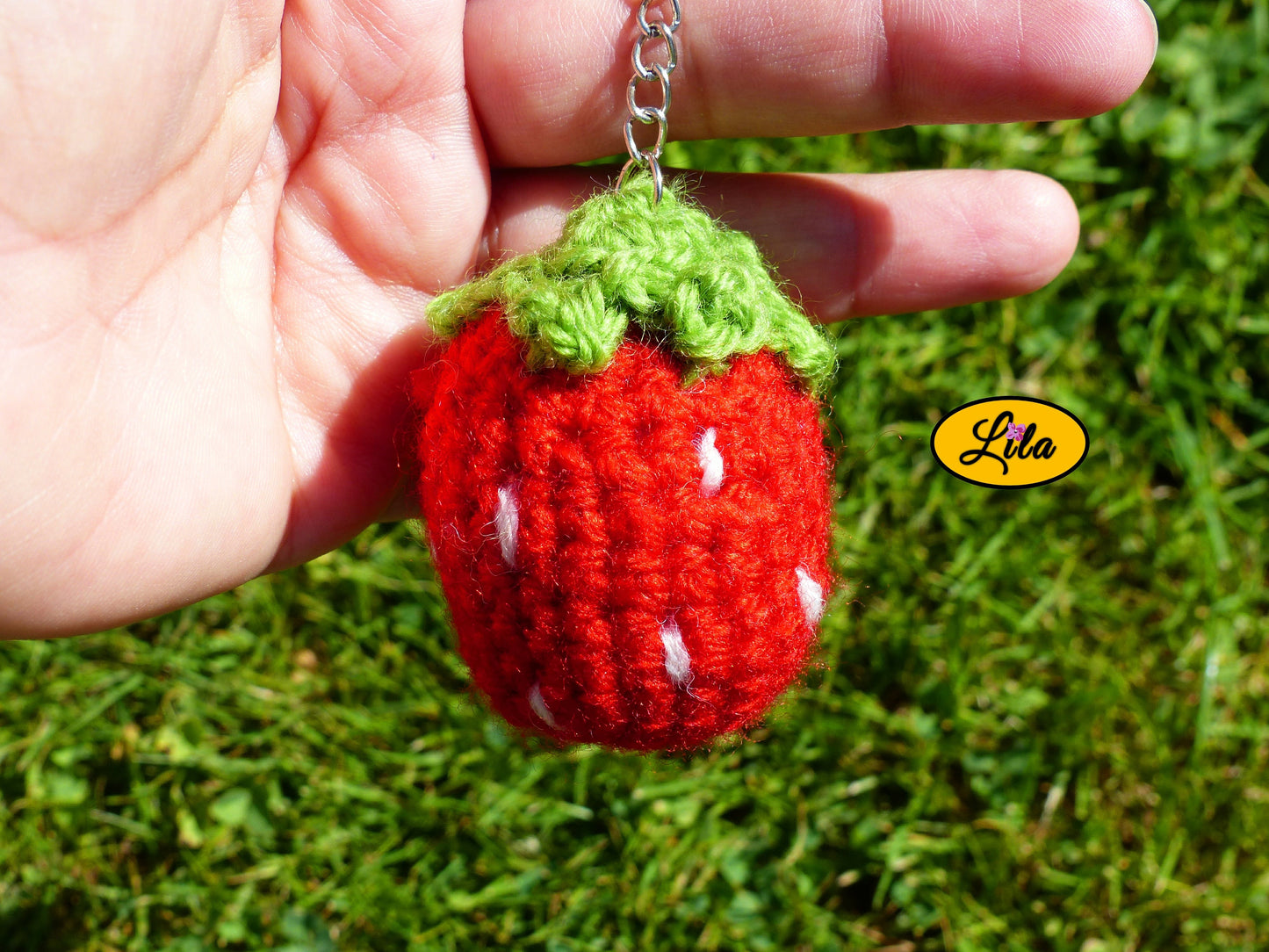 Porte-clé fraise amigurumi rouge avec points blancs et queue vert clair tenu dans une main devant une pelouse style artisanal et gourmand
