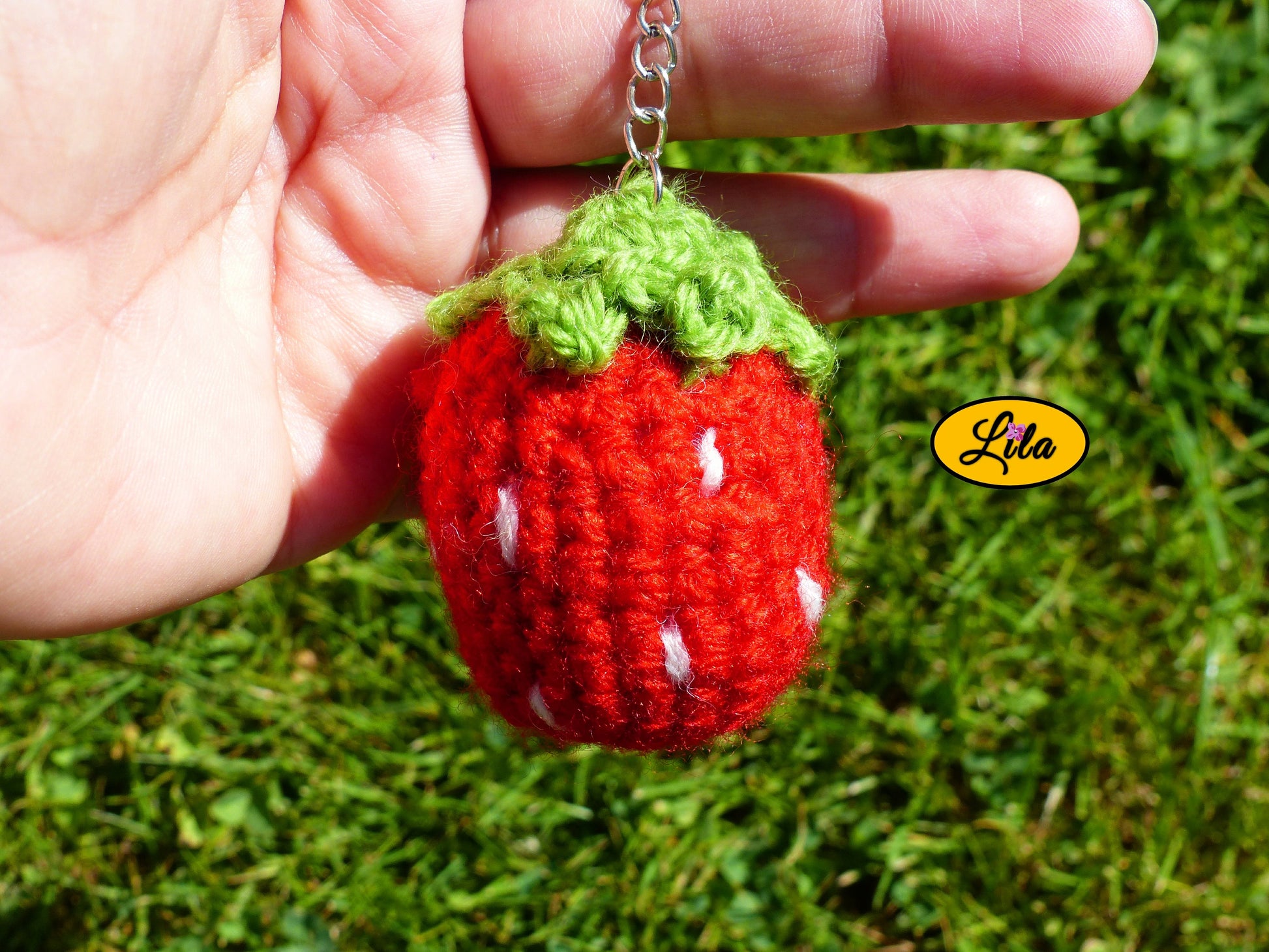 Porte-clé fraise amigurumi rouge avec points blancs et queue vert clair tenu dans une main devant une pelouse style artisanal et gourmand