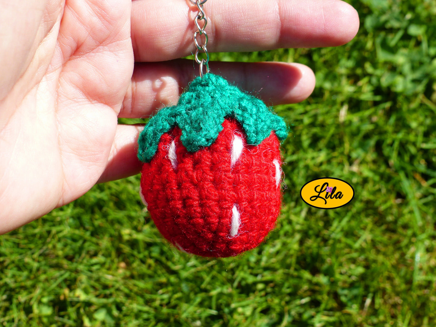 Porte-clé fraise amigurumi rouge avec points blancs et queue vert foncé tenu dans une main avec fond pelouse style kawaii et crochet handmade