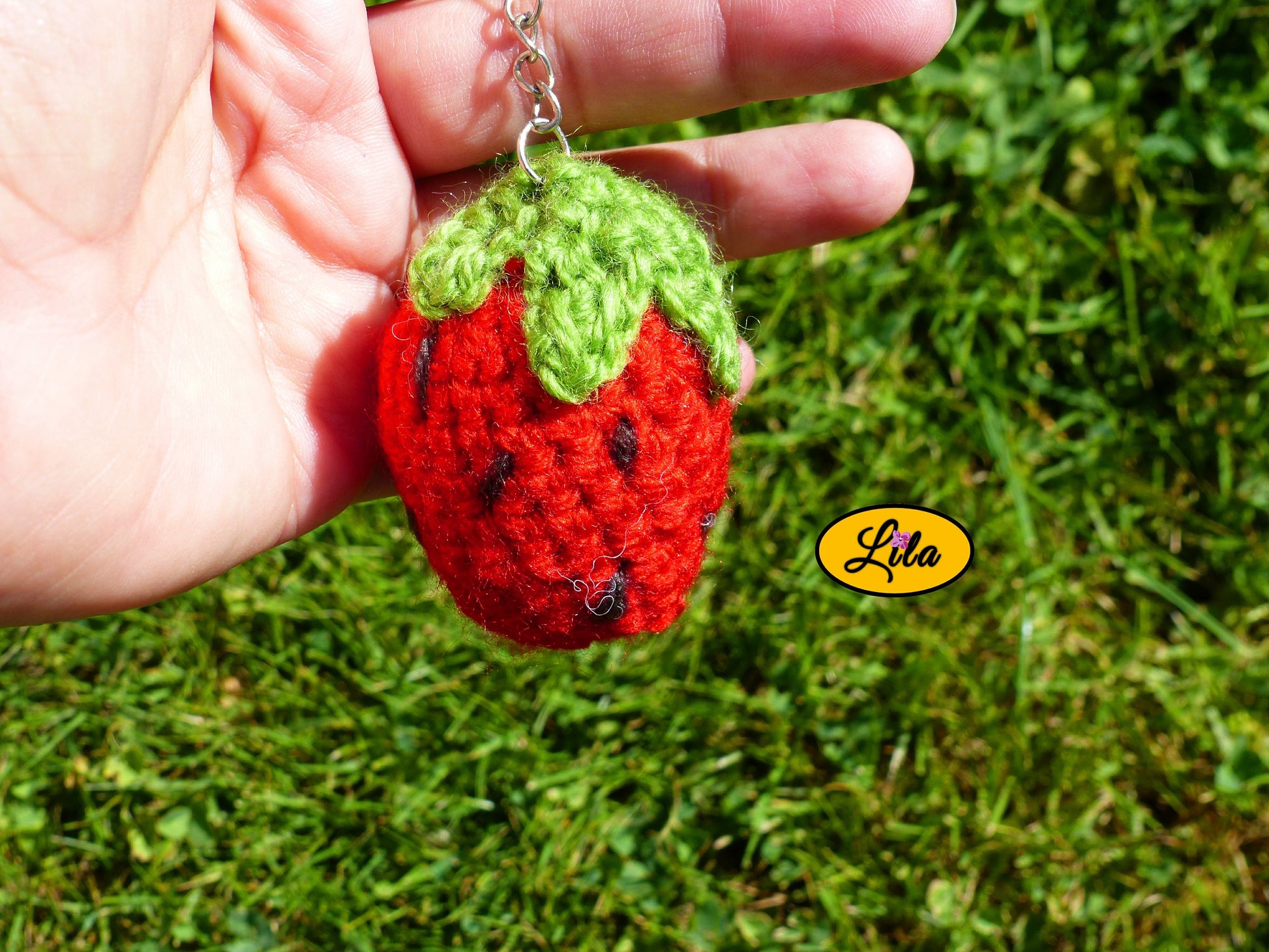 Porte-clé fraise amigurumi rouge avec points noirs et queue vert clair tenu dans une main avec pelouse en arrière-plan style handmade et kawaii