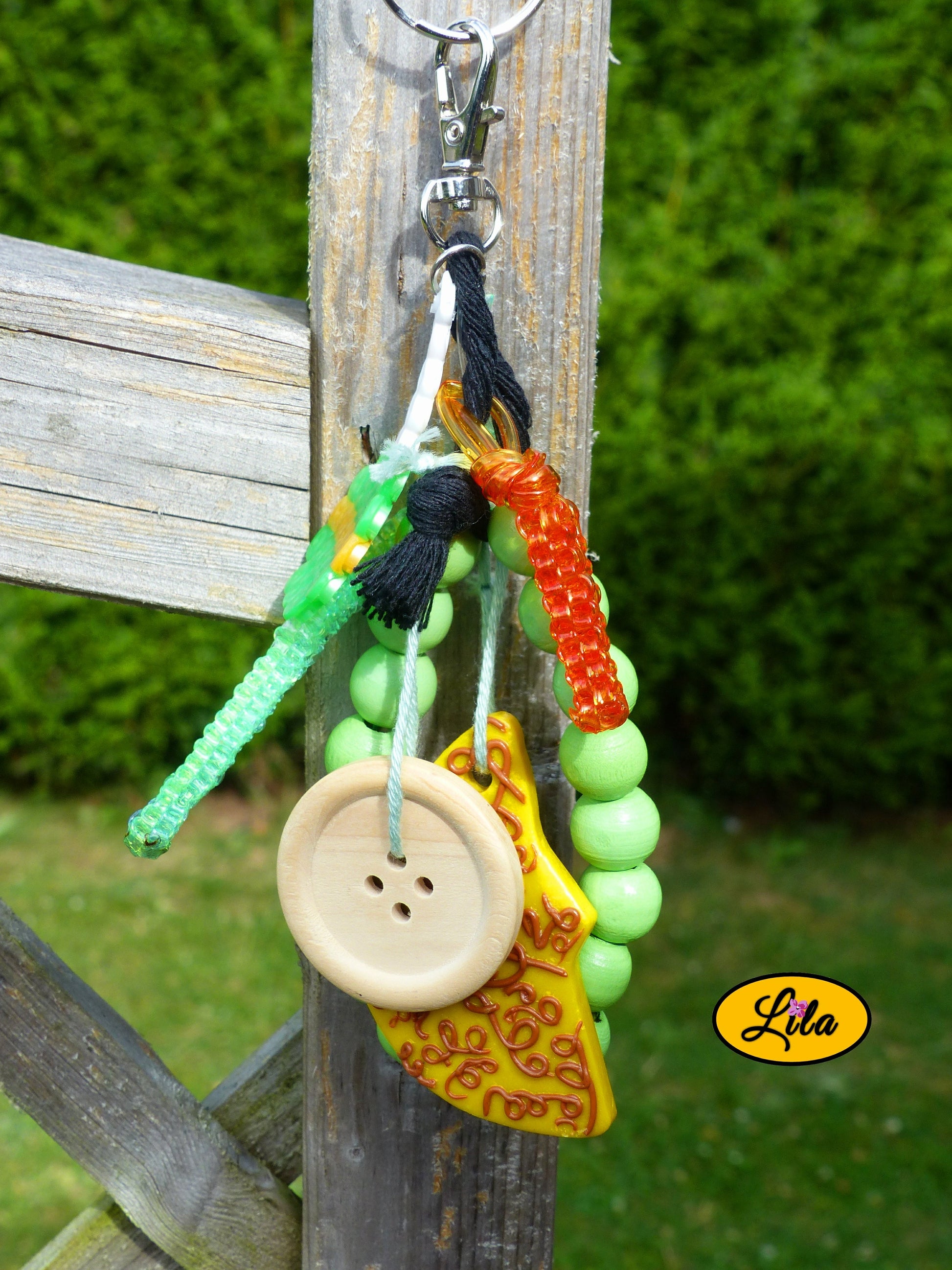 Porte-clés lune dorée esprit nature composé d’une sucette verte en perles à repasser avec bâtonnet blanc, lune jaune ornée de motifs bruns en pâte polymère, bracelet en grosses perles vertes, scoubidous colorés, bouton en bois et bouts de laine tressée suspendus à un mousqueton style bohème et artisanal