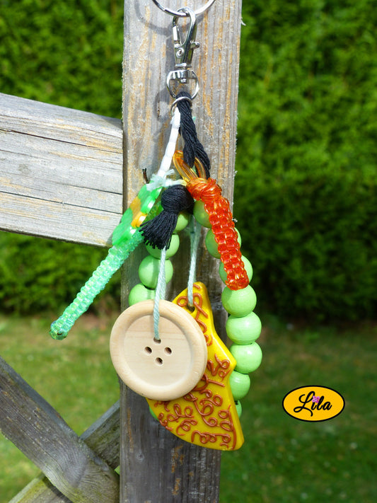 Porte-clés lune dorée esprit nature composé d’une sucette verte en perles à repasser avec bâtonnet blanc, lune jaune ornée de motifs bruns en pâte polymère, bracelet en grosses perles vertes, scoubidous colorés, bouton en bois et bouts de laine tressée suspendus à un mousqueton style bohème et artisanal
