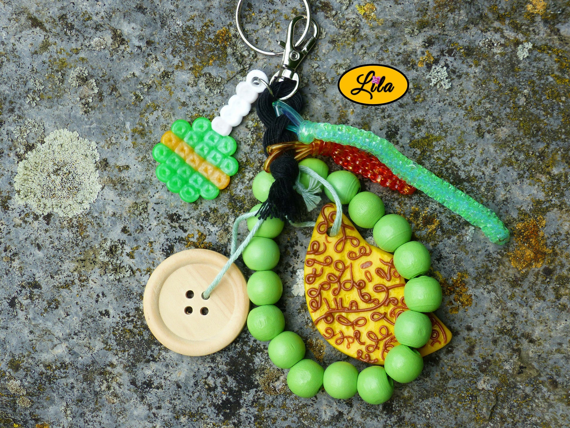 Porte-clés lune dorée esprit nature posé sur une pierre vue de haut avec sucette en perles à repasser verte, lune décorée en pâte polymère, grosses perles vertes, bouton en bois et scoubidous visibles côte à côte style créatif et récup