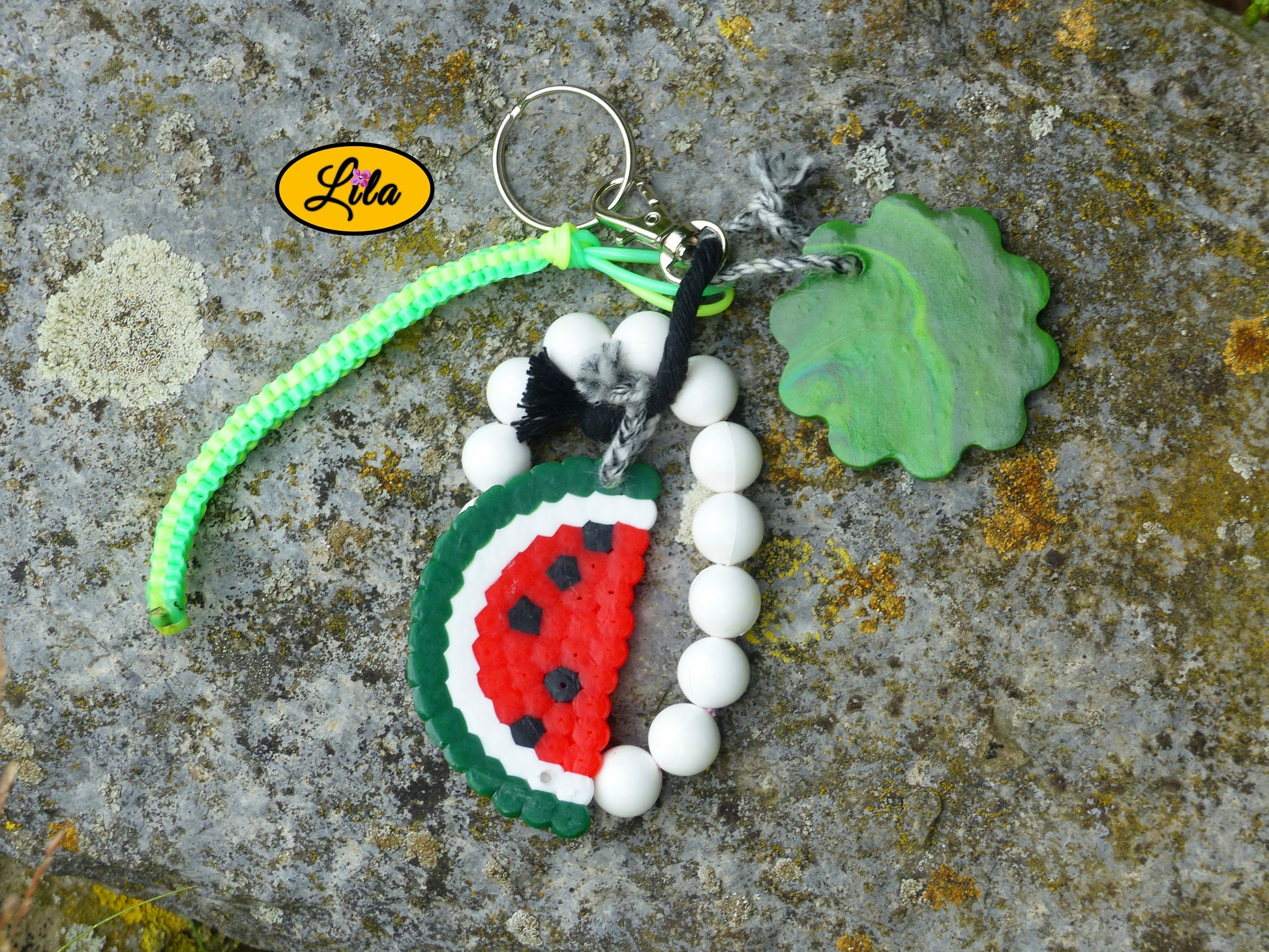 Porte-clés pastèque et perles posé sur une pierre vue de haut avec demi-tranche de pastèque en perles à repasser, fleur verte en pâte polymère, grosses perles blanches et scoubidou visibles côte à côte style créatif et fruité