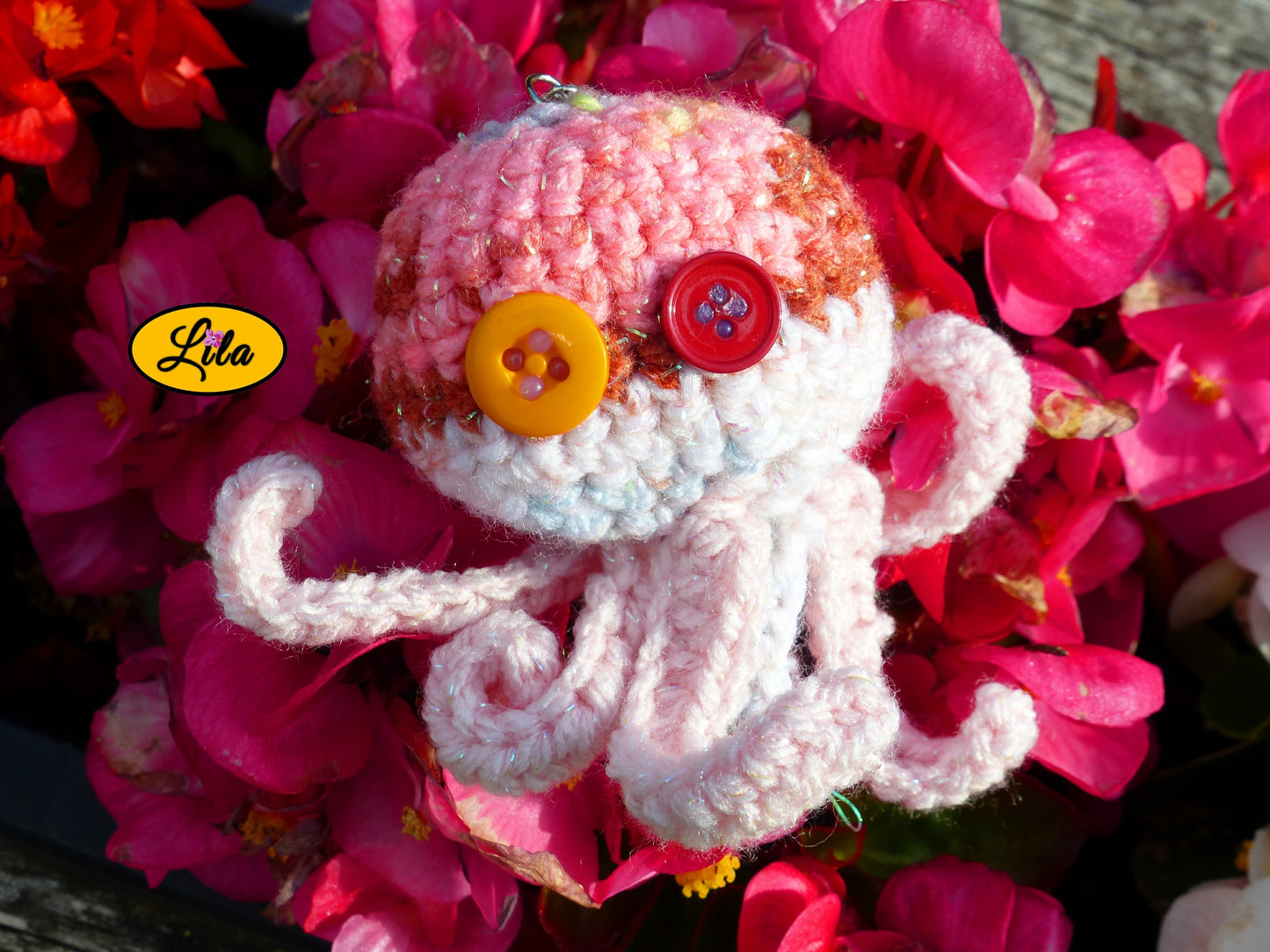 Porte-clé Poulpy pieuvre amigurumi rose avec dégradés pailletés réalisée au crochet avec yeux en boutons orange et fuchsia posé sur un lit de fleurs style kawaii et artisanal