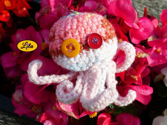 Porte-clé Poulpy pieuvre amigurumi rose avec dégradés pailletés réalisée au crochet avec yeux en boutons orange et fuchsia posé sur un lit de fleurs style kawaii et artisanal