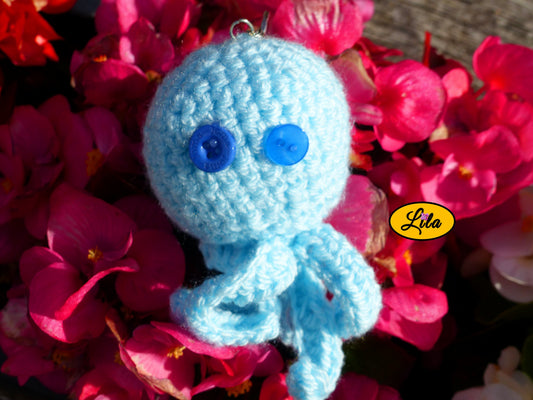 Porte-clé Poulpy pieuvre amigurumi bleu ciel réalisée au crochet avec yeux en boutons bleus de différentes épaisseurs posé sur un lit de fleurs style kawaii et artisanal
