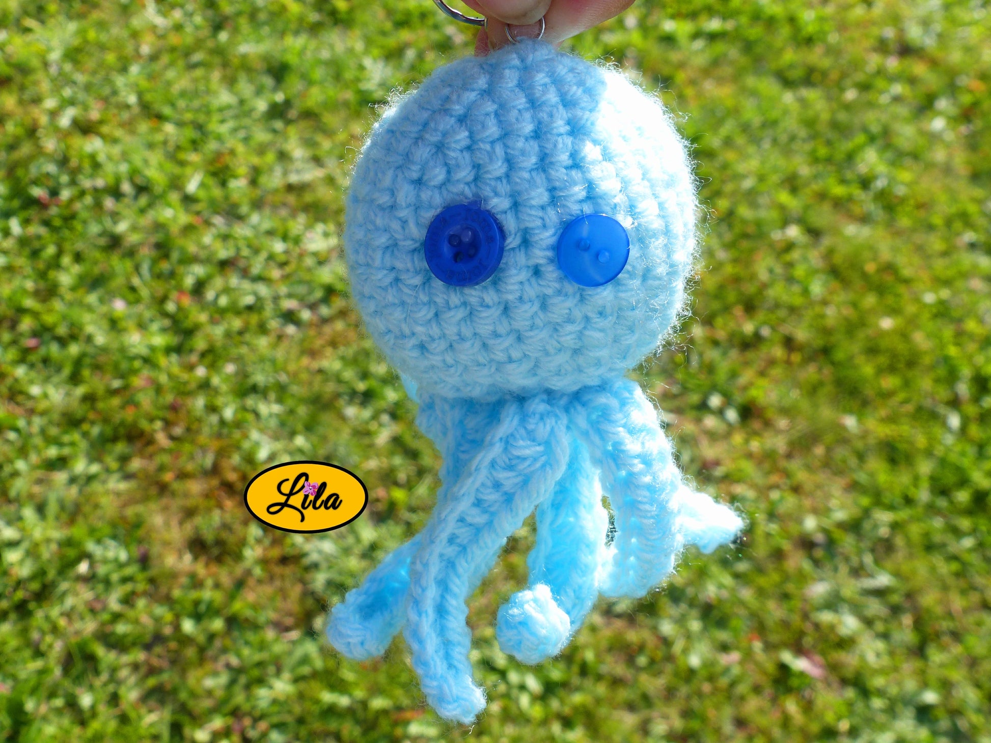 Porte-clé Poulpy pieuvre amigurumi bleu ciel réalisée au crochet avec yeux en boutons bleus de différentes épaisseurs tenu dans une main en extérieur dans un jardin style handmade et mignon