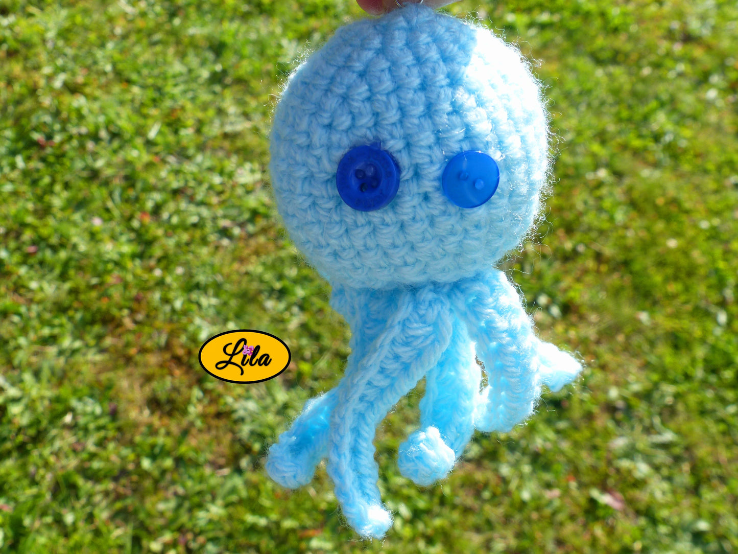 Photo rapprochée du porte-clé Poulpy pieuvre amigurumi bleu ciel au crochet avec yeux en boutons bleus de différentes tailles dans un jardin style artisanal et kawaii
