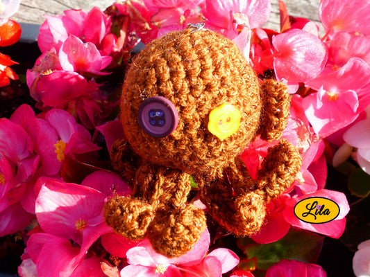 Porte-clé Poulpy pieuvre amigurumi brune pailletée réalisée au crochet avec yeux en boutons fleur jaune et mauve foncé posé sur un lit de fleurs style bohème et artisanal