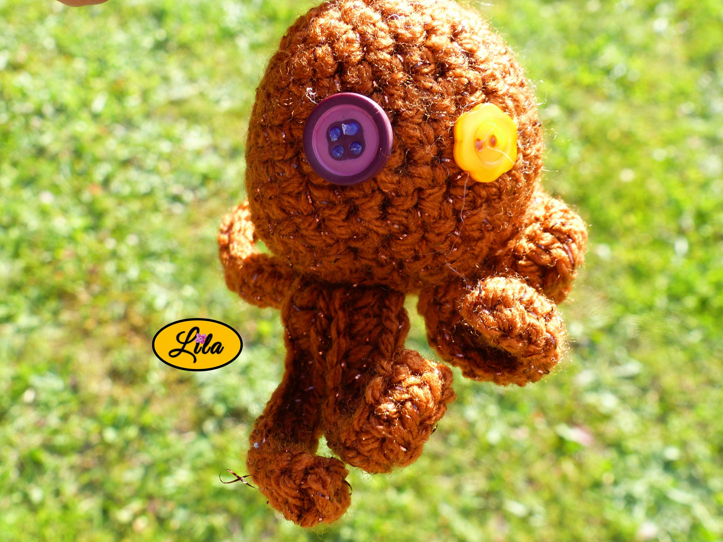 Photo rapprochée du porte-clé Poulpy pieuvre amigurumi brune pailletée au crochet avec yeux en boutons fleur jaune et mauve foncé dans un jardin style artisanal et kawaii
