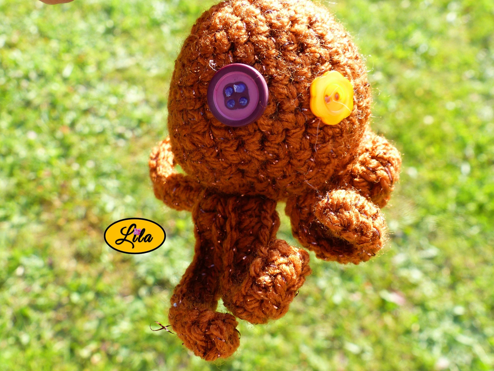 Photo rapprochée du porte-clé Poulpy pieuvre amigurumi brune pailletée au crochet avec yeux en boutons fleur jaune et mauve foncé dans un jardin style artisanal et kawaii