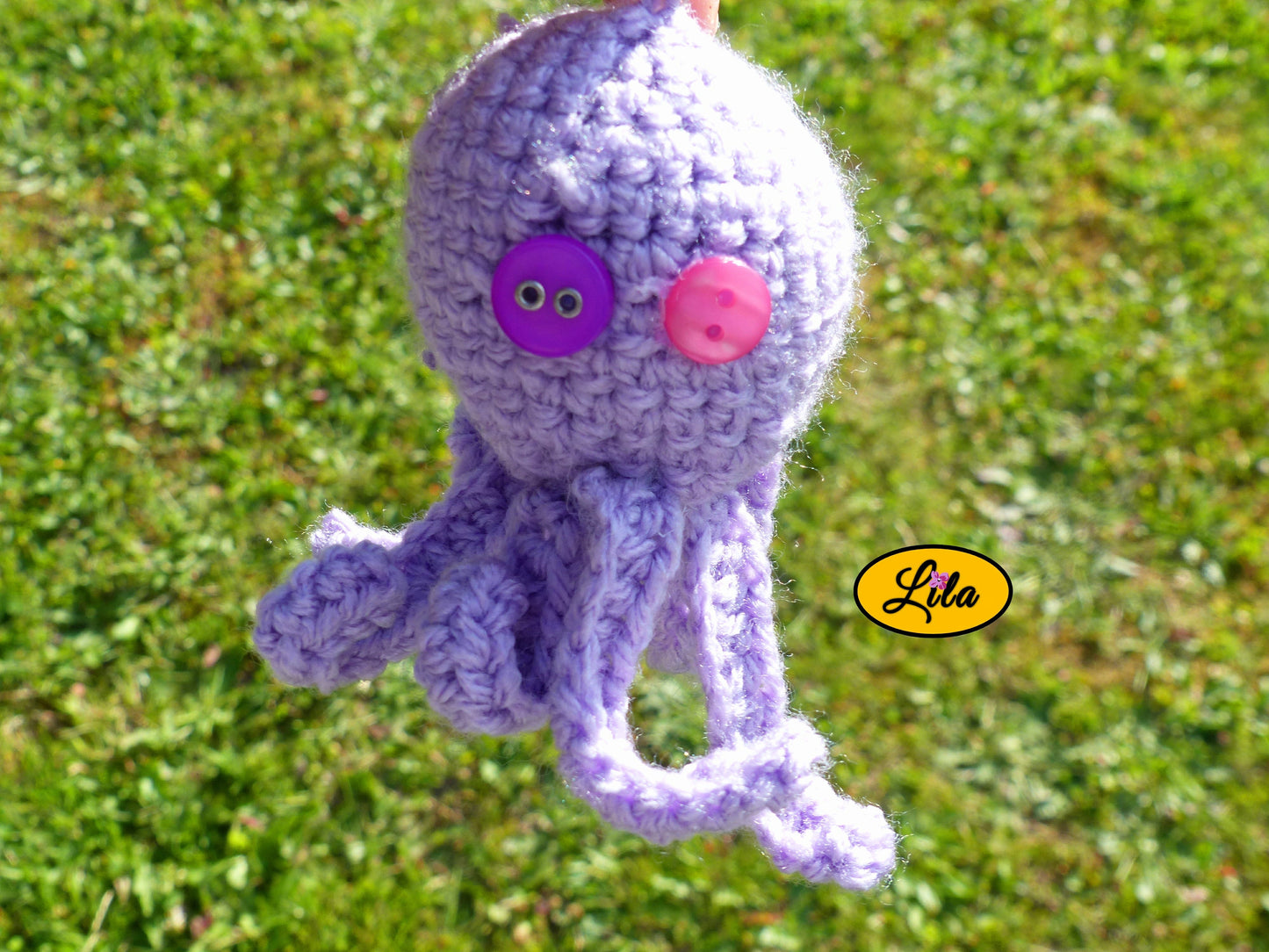 Photo rapprochée du porte-clé Poulpy pieuvre amigurumi mauve clair au crochet avec yeux en boutons mauve et rose dans un jardin style artisanal et kawaii
