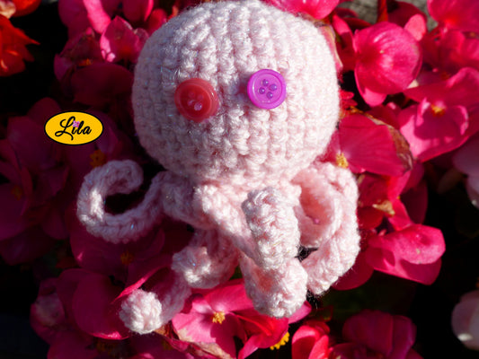 Porte-clé Poulpy pieuvre amigurumi rose clair réalisée au crochet avec yeux en boutons rose et mauve posé sur un lit de fleurs roses style kawaii et artisanal