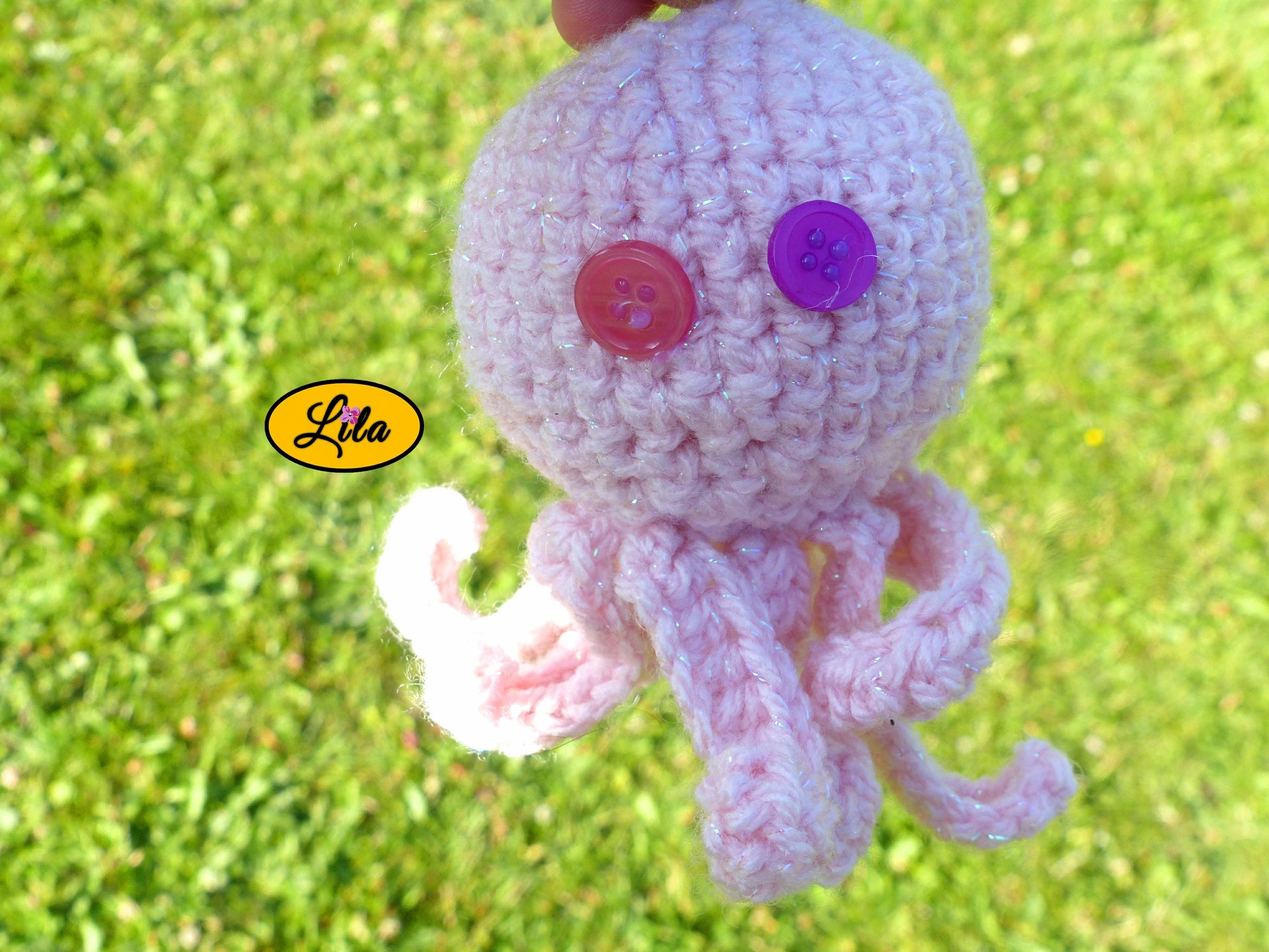 Porte-clé Poulpy pieuvre amigurumi rose clair réalisée au crochet photographié sous un autre angle dans un jardin avec yeux en boutons dépareillés rose et mauve style kawaii et bohème