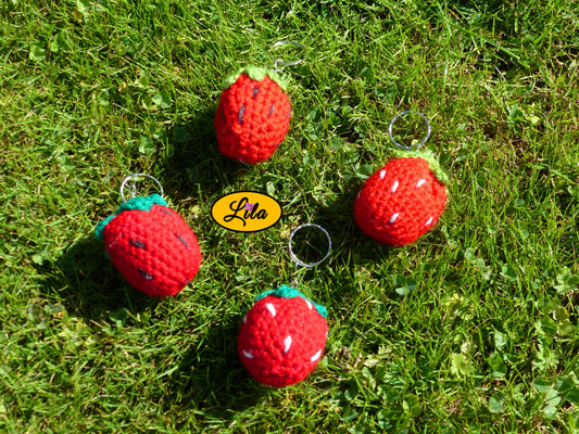 Quatre porte-clés fraises amigurumi réalisés au crochet avec différentes queues vert clair ou vert foncé et graines blanches ou noires posés sur une pelouse style kawaii et artisanal