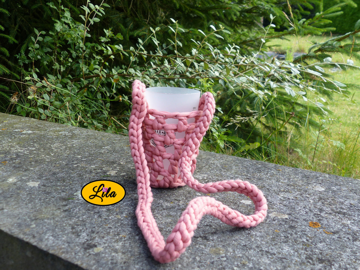 Porte-gobelet rose avec tresse pour port autour du cou façon collier pendant et gobelet à l’intérieur posé de face sur une pierre en extérieur style festival et artisanal