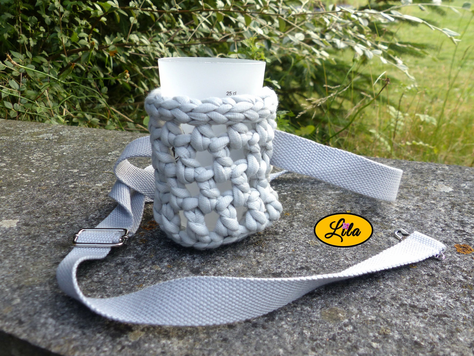 Porte-gobelet crocheté gris avec ceinture grise et gobelet à l’intérieur posé de face sur une pierre en extérieur style festival et artisanal