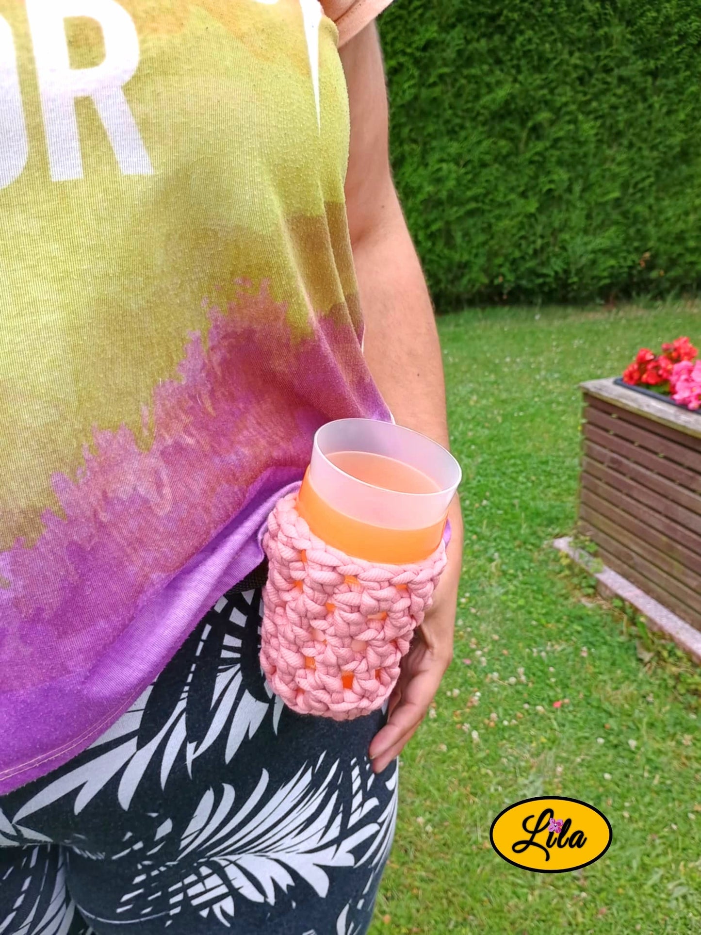 Porte-gobelet crocheté rose avec ceinture noire porté de face par un mannequin dans un jardin avec un gobelet à l’intérieur style bohème et festival
