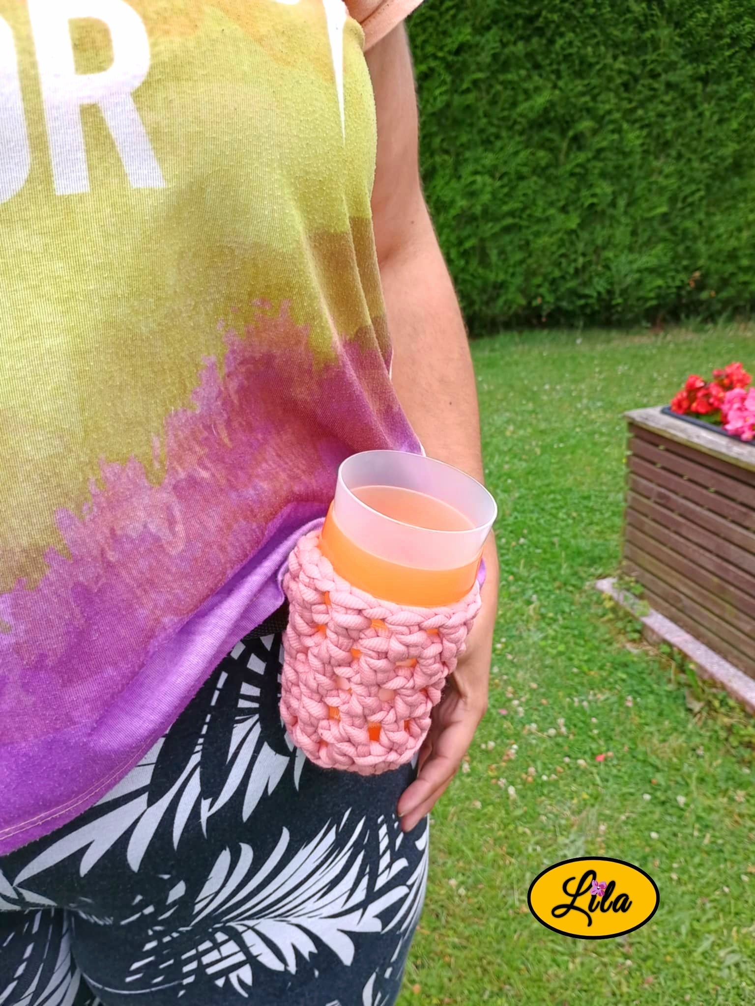 Porte-gobelet crocheté rose avec ceinture noire porté de face par un mannequin dans un jardin avec un gobelet à l’intérieur style bohème et festival