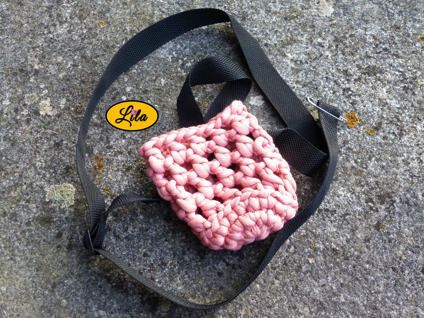 Porte-gobelet crocheté en laine rose avec ceinture noire vu de haut à plat sur une pierre en extérieur sans gobelet style festival et artisanal