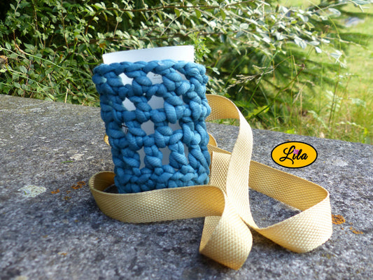 Porte-gobelet crocheté en laine turquoise avec ceinture couleur sable et gobelet posé de face sur une pierre en extérieur style bohème et artisanal