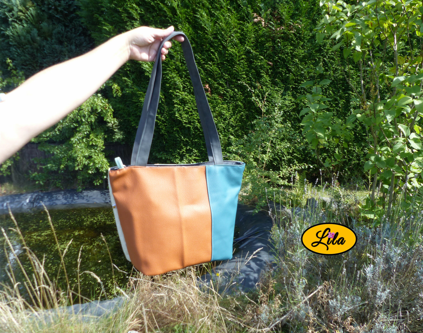 Sac 4 saisons en simili cuir brun et turquoise porté à la main devant un jardin et un étang style moderne et coloré